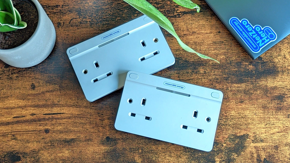 Two measurable.energy double gang plug sockets on a dark wood table, alongisde a plant and laptop.